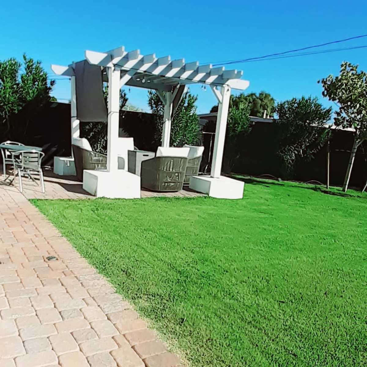 A white pergola with seating is set on a green lawn next to a stone pathway.