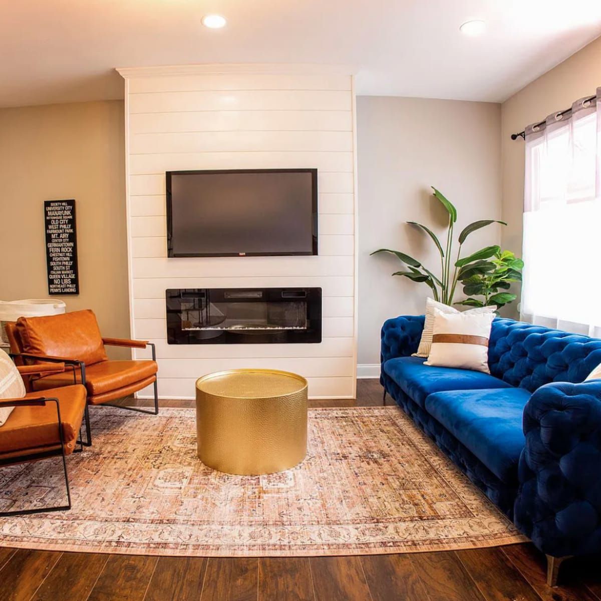 The living room features a blue upholstered sofa, two brown leather chairs, a gold coffee table, and a wall-mounted television above an electric fireplace, all arranged on a patterned area rug.