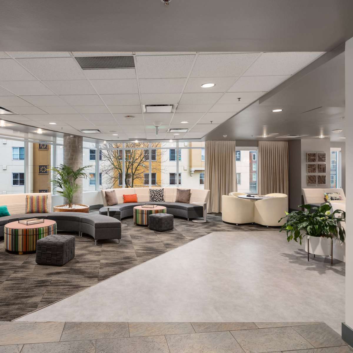 A modern lounge area with a curved sectional sofa, colorful cushions, small tables, and large windows allowing natural light to fill the space.