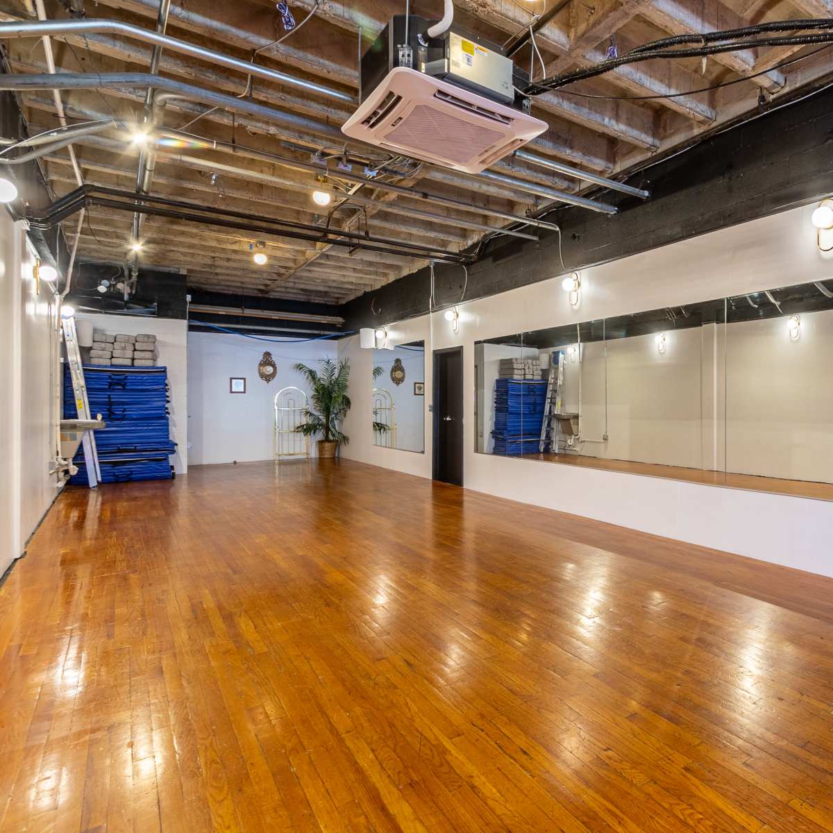 An empty studio space with wooden floors, a large mirror on one wall, and exposed beams on the ceiling.