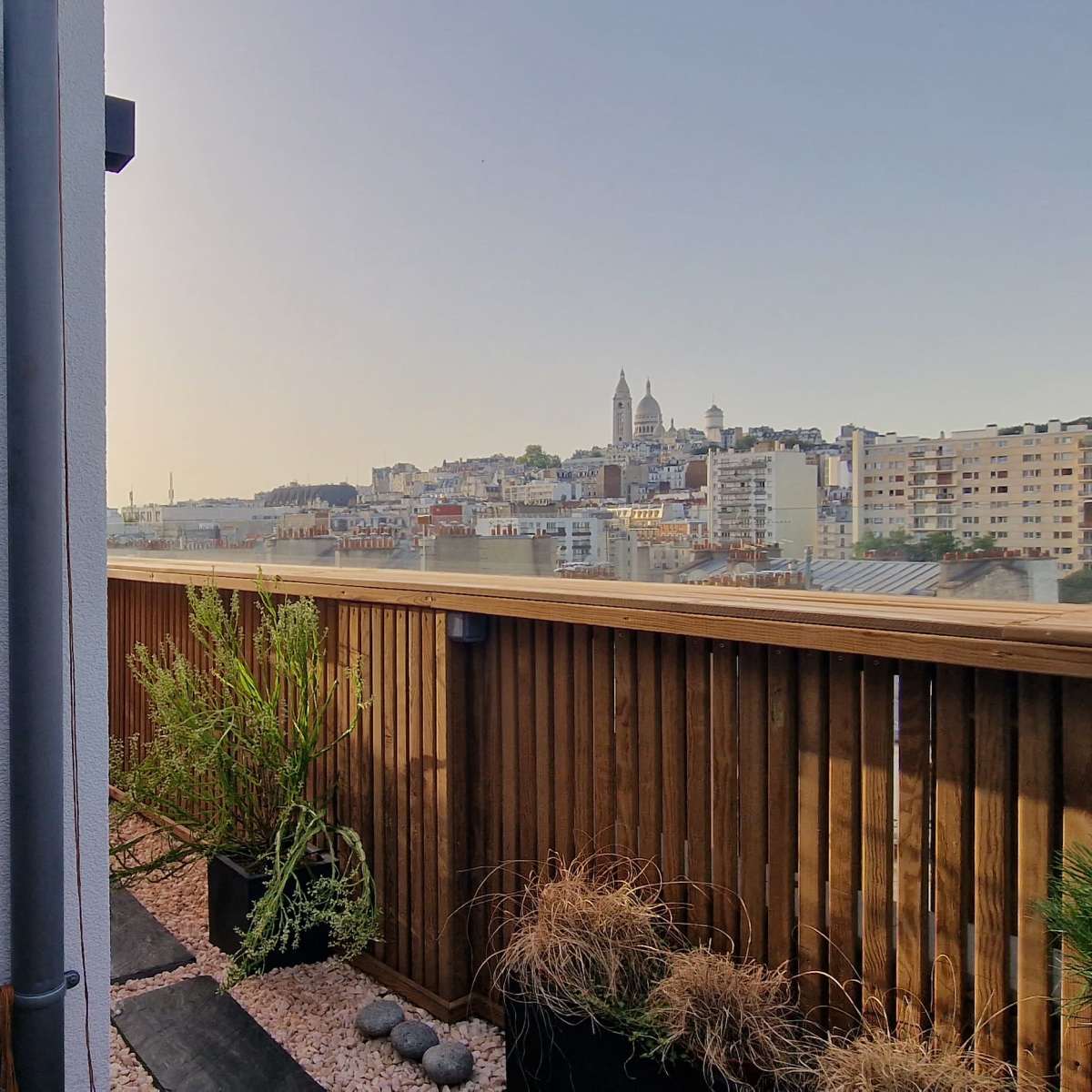 A view from a terrace shows a cityscape with buildings and a distant church spire under a clear sky.