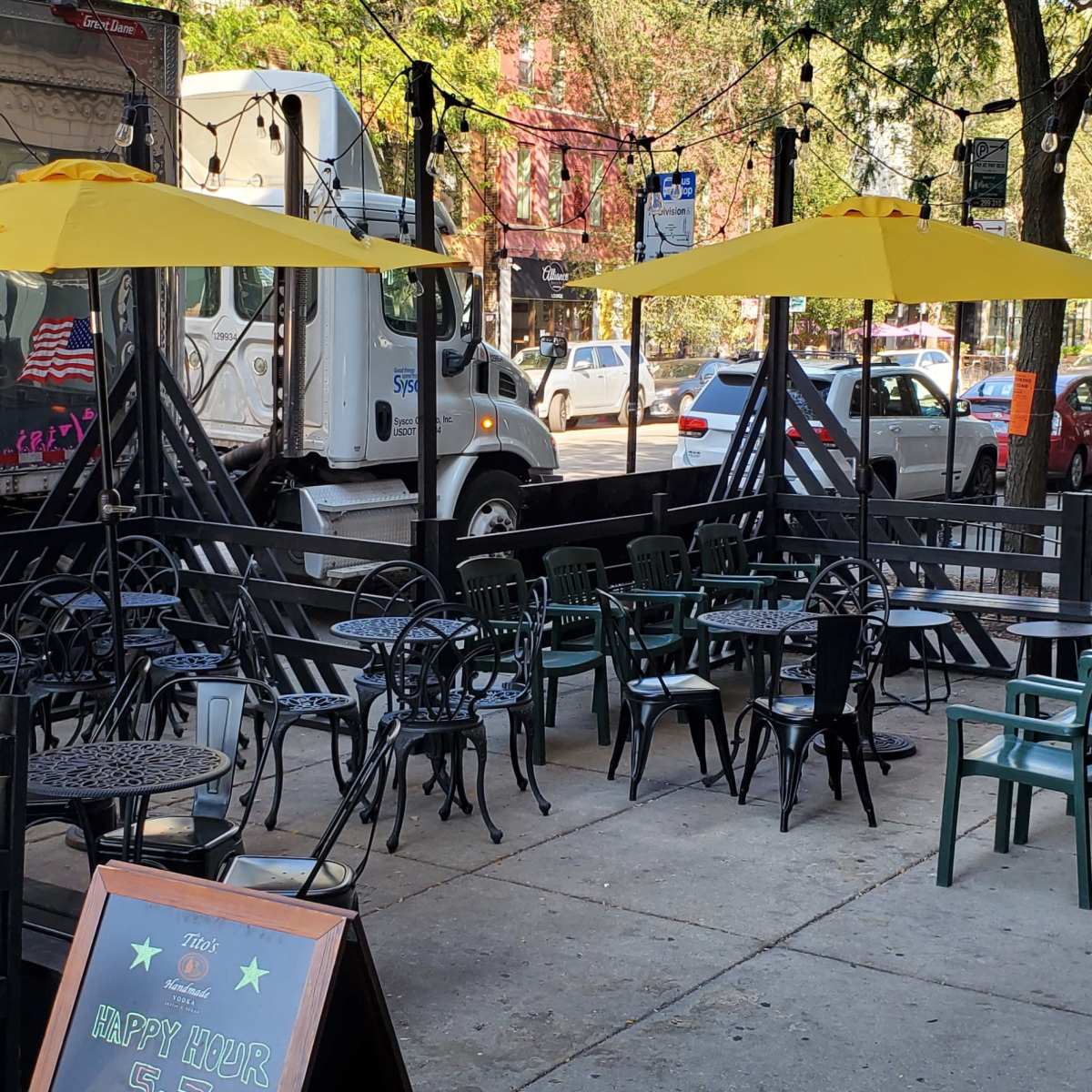 A street-side outdoor dining area features metal tables and chairs shaded by yellow umbrellas, adjacent to a parked delivery truck.