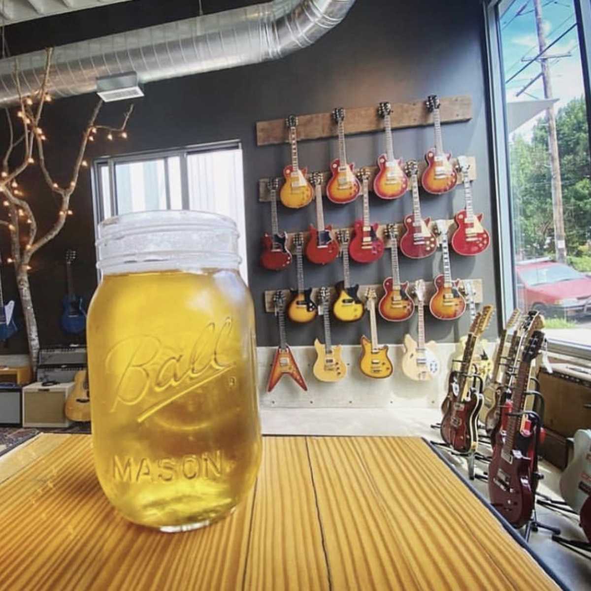 A mason jar filled with a light-colored beverage sits on a wooden table in a room with various electric guitars displayed on the walls.