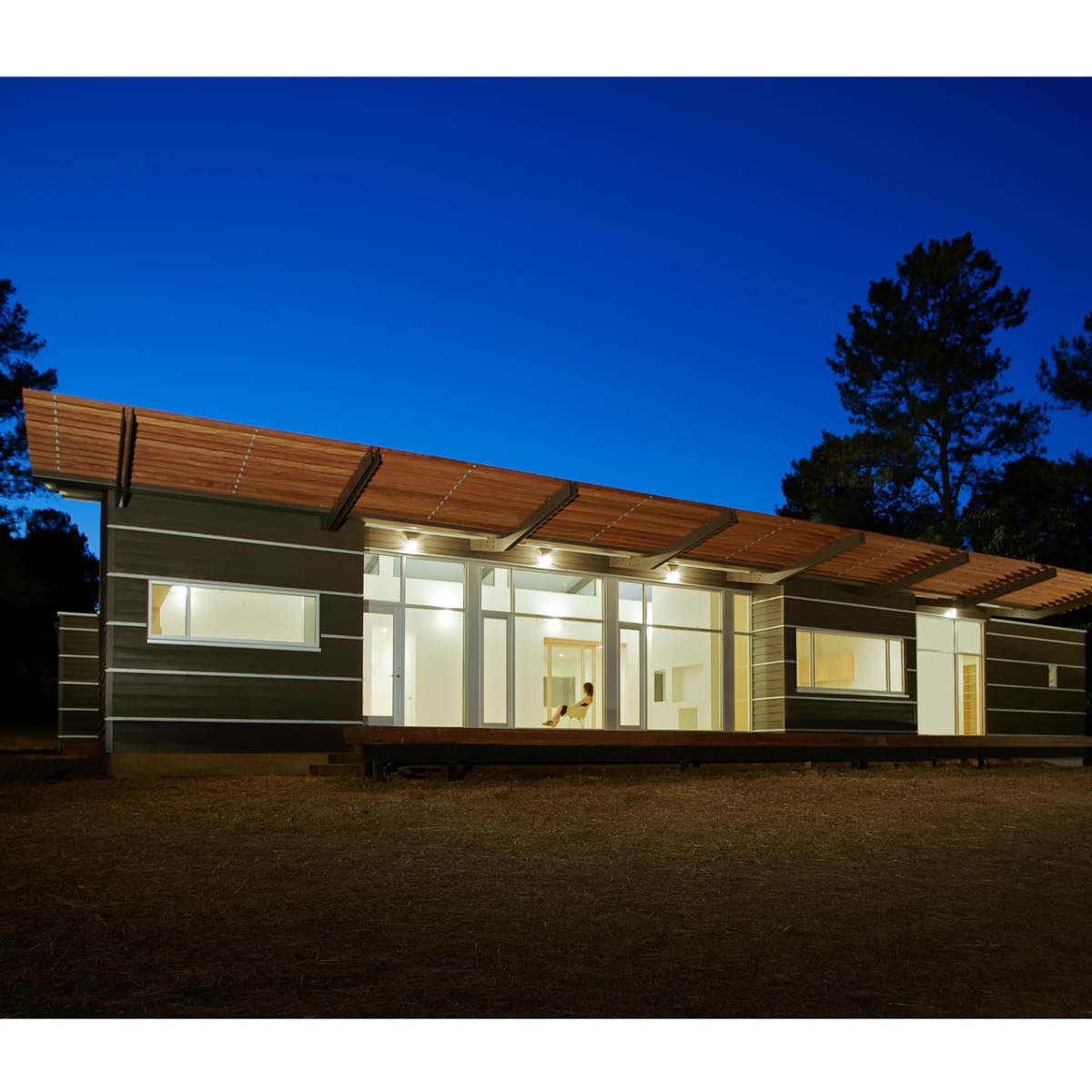 A modern, single-story house with a wooden overhang is illuminated against a deep blue evening sky, surrounded by tall trees.