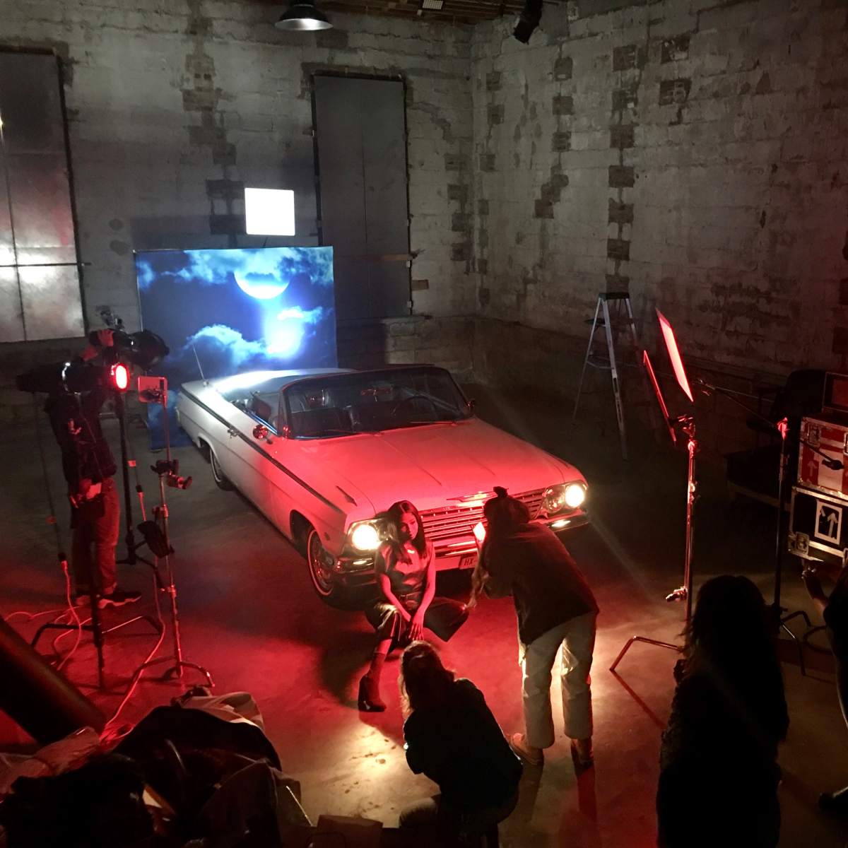 A film crew sets up lighting and equipment around a classic car in a dimly lit industrial space.