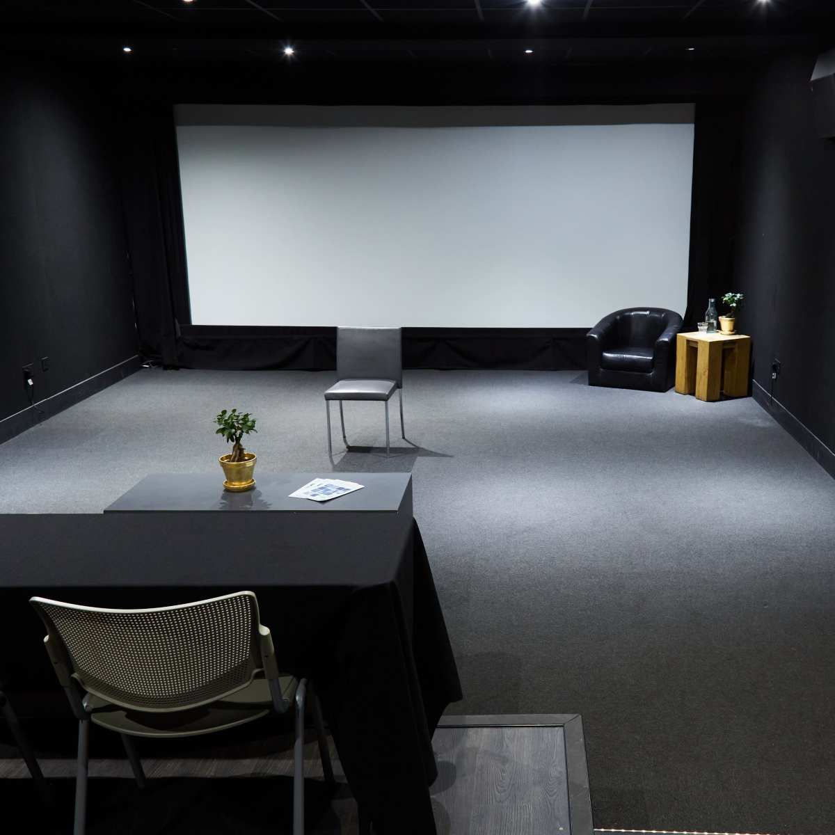 A sparsely furnished black walled room with a large projection screen, a single chair, a small table, and a black armchair beside a wooden side table.