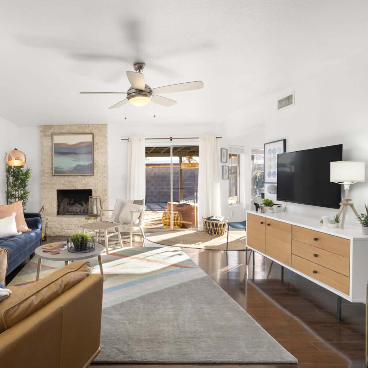 A bright living room features a blue sofa, a tan leather chair, a television unit, and a fireplace with large windows allowing natural light.