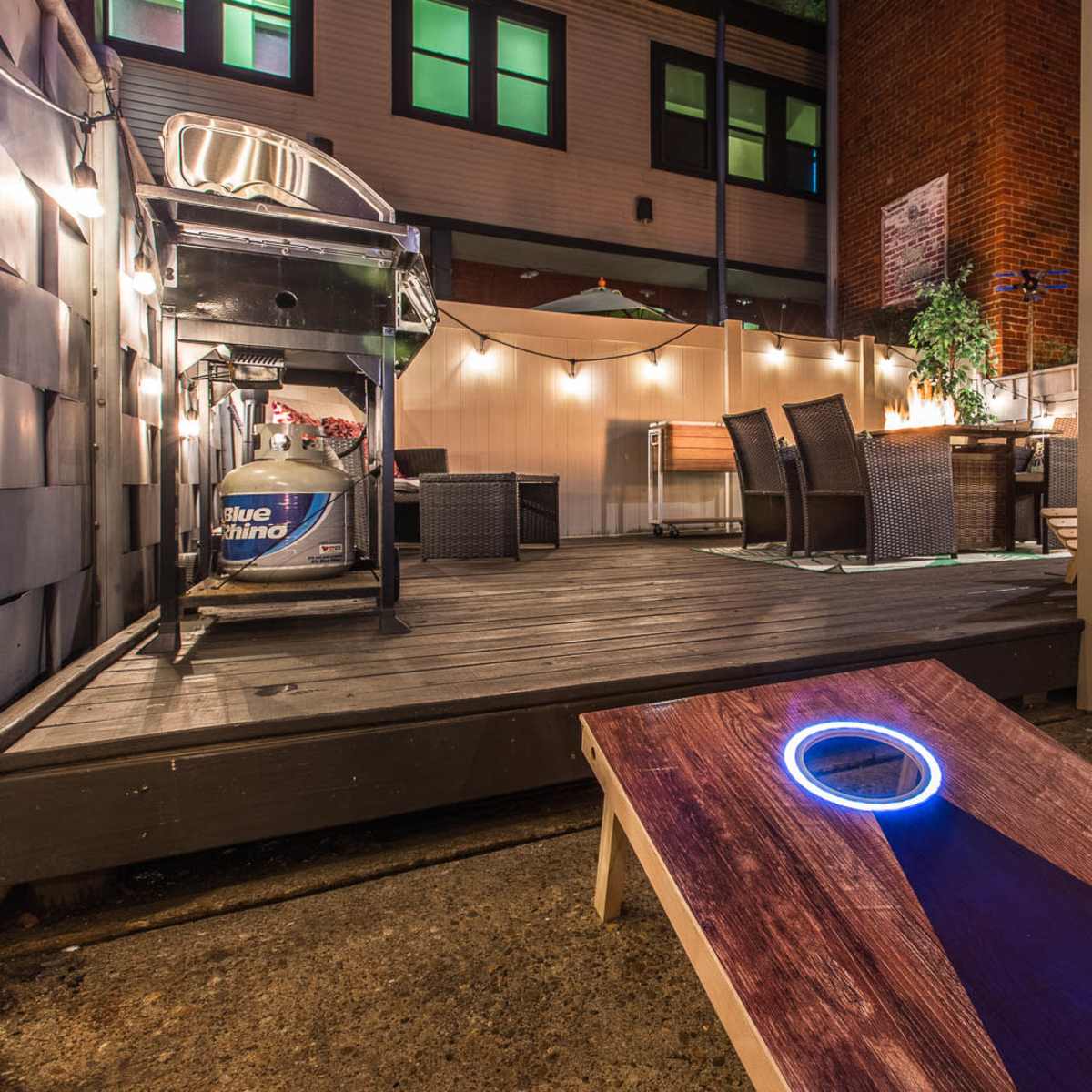 A backyard space with a cornhole game setup, a grill, outdoor dining furniture, and ambient lighting.