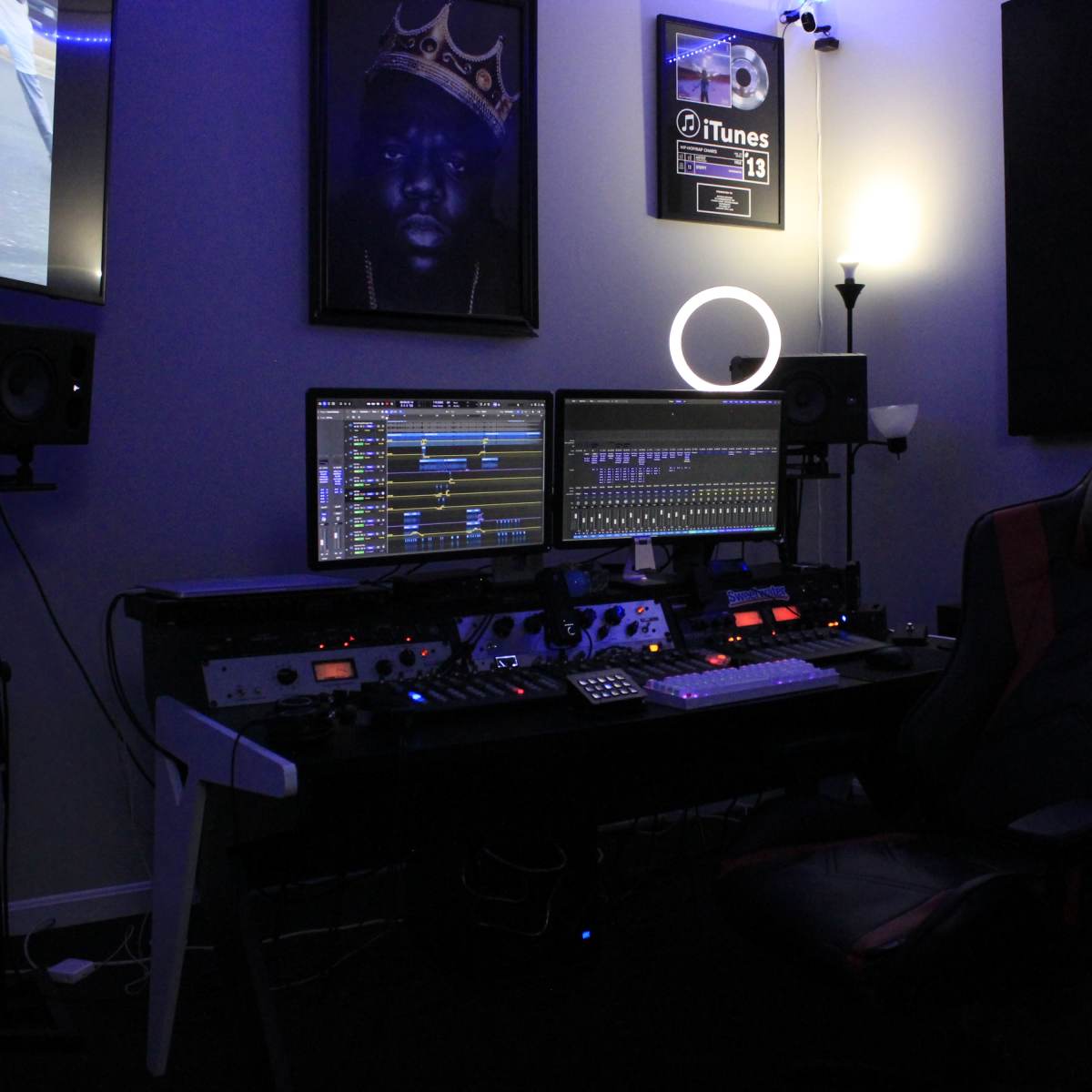 A music production studio with two computer monitors, audio equipment, and a gaming chair under dim lighting.