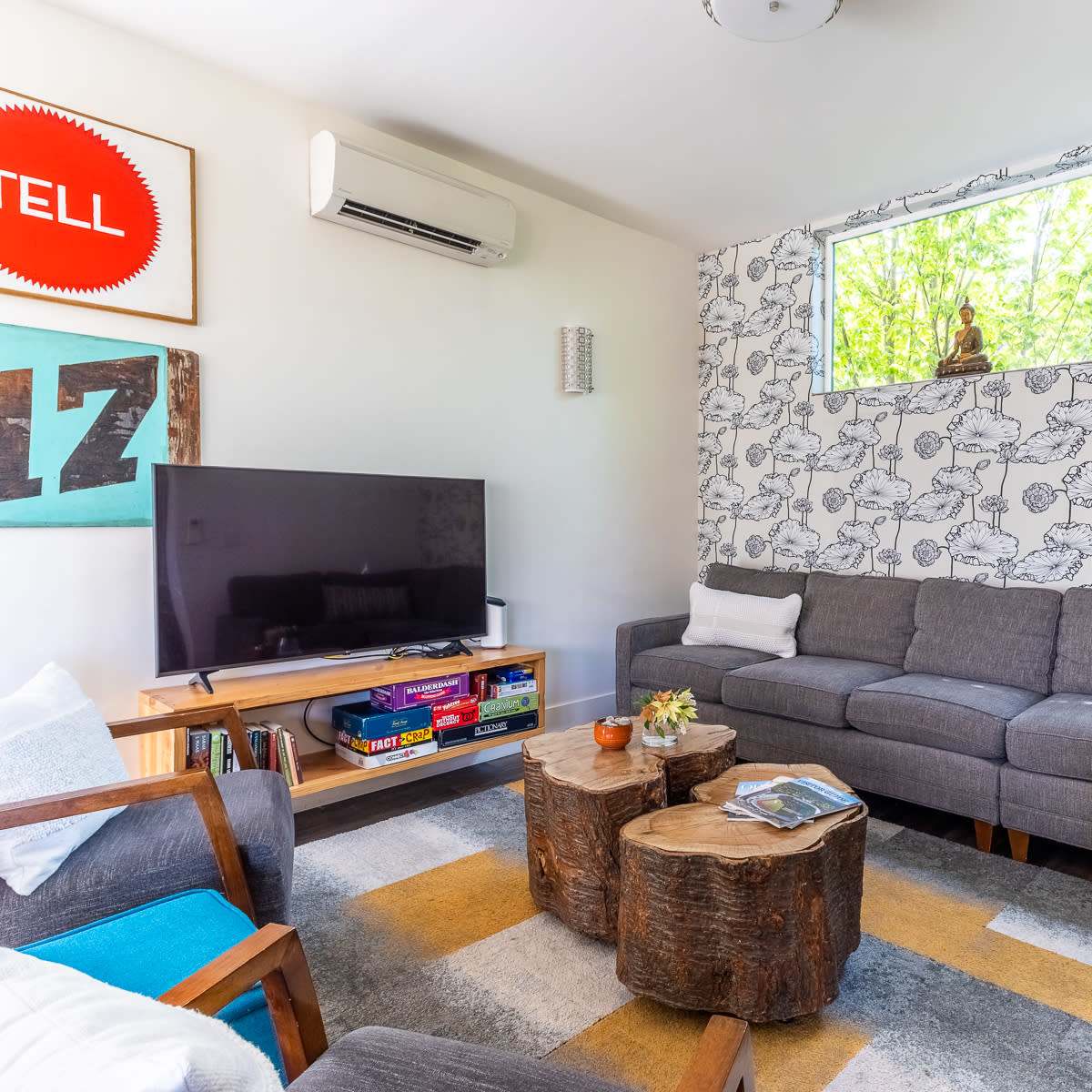 A modern living room with a gray sectional sofa, a wooden coffee table made from tree stumps, a television on a wooden stand, and large windows allowing natural light to enter.