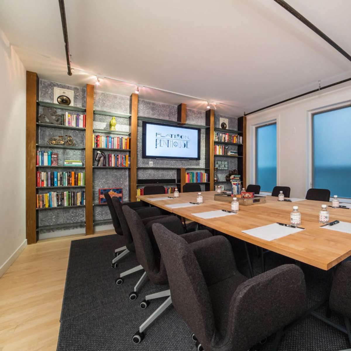 A modern conference room with a large wooden table surrounded by black chairs, a TV screen on the wall, and bookshelves filled with books.