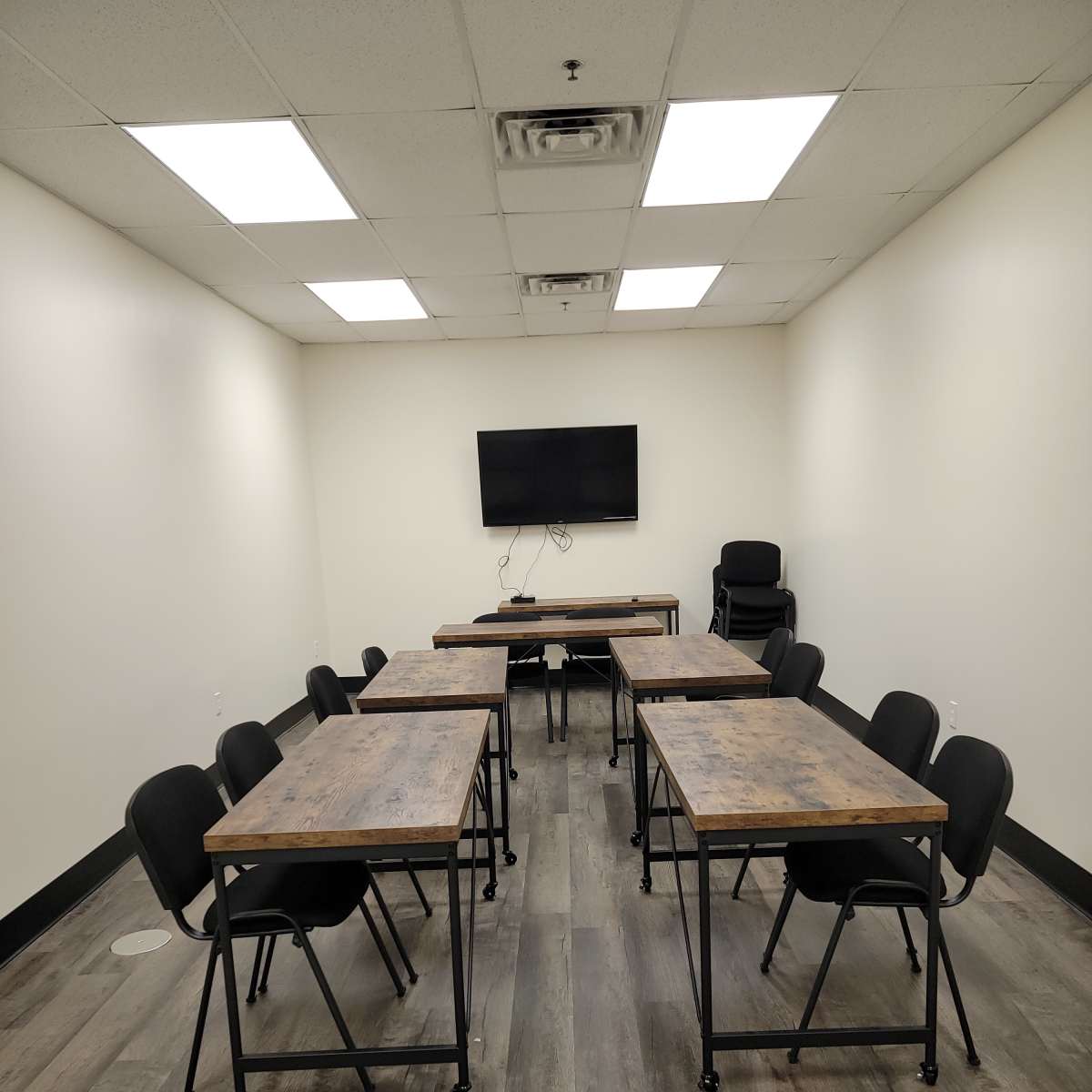 A meeting room is arranged with several wooden tables and black chairs, featuring a wall-mounted television.