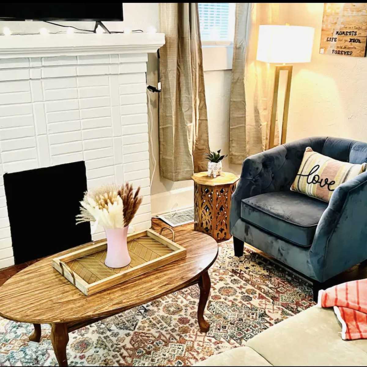 A cozy living room featuring a blue armchair with a "love" pillow, a wooden coffee table with a tray and a vase of dried flowers, and a patterned area rug.