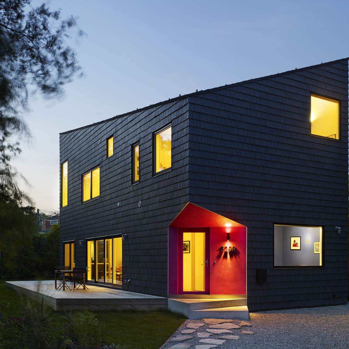 A modern two-story house with a dark shingle exterior and a prominently colored entryway, situated in a landscaped yard at dusk.