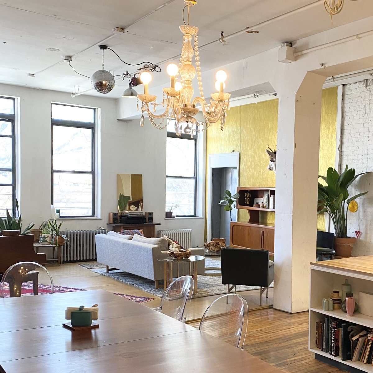 An open, airy loft space with large windows, a dining table, a chandelier, and a cozy living area featuring plants and modern furniture.