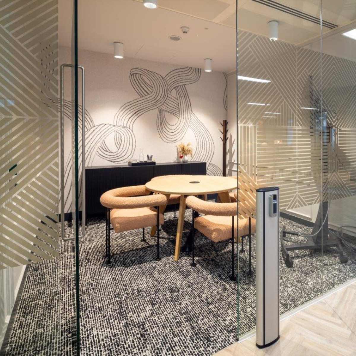 A modern office space with a glass-walled meeting room featuring a round table and stylish chairs, complemented by patterned walls and wooden flooring.