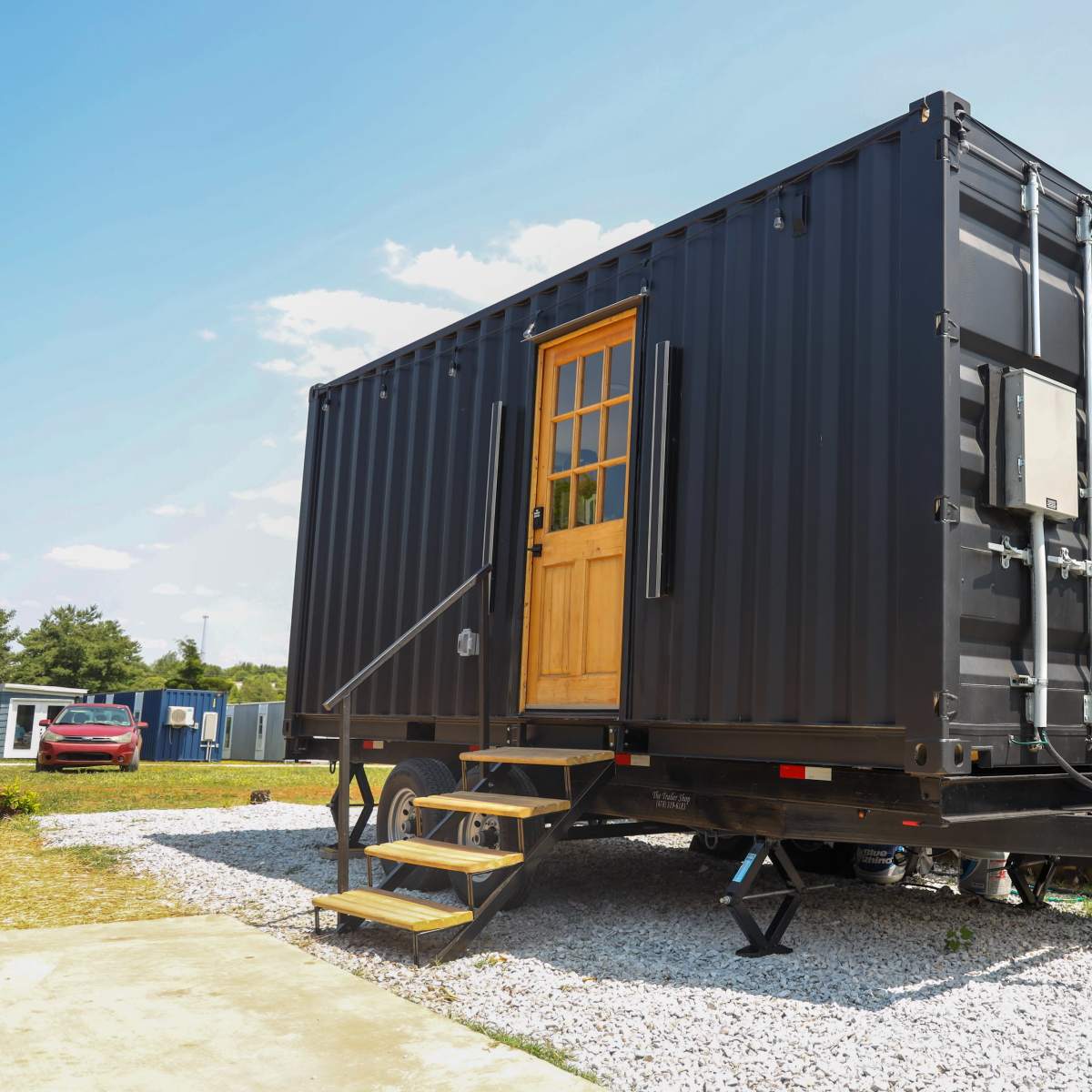 A black shipping container is set on a trailer with a wooden door and air conditioning unit, surrounded by gravel and a wooden fence.