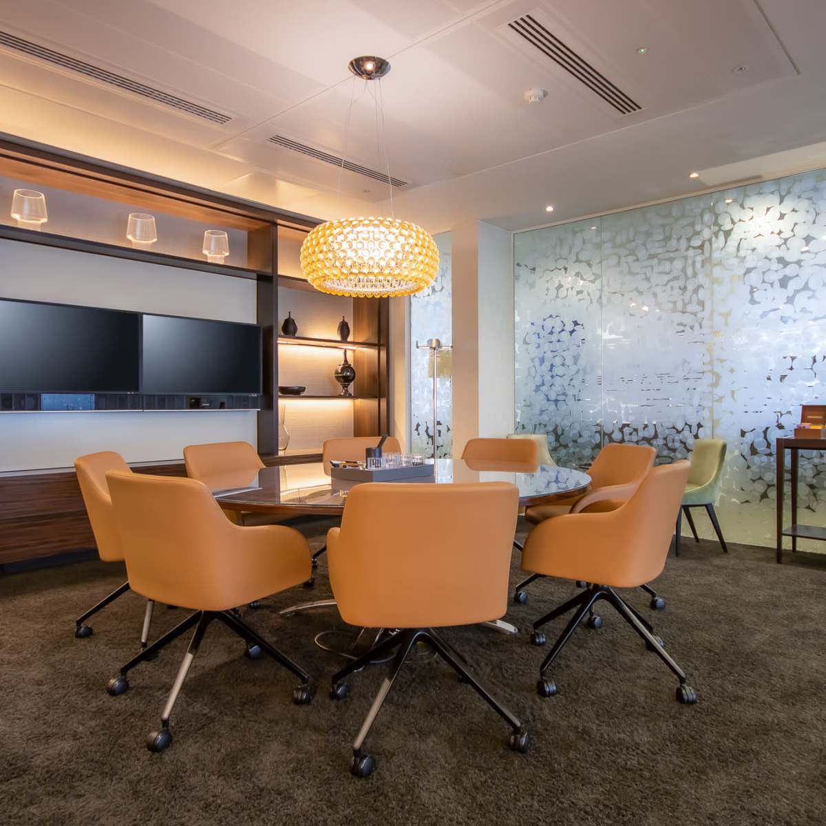 A modern meeting room featuring a round table surrounded by orange chairs, a large wall-mounted screen, and decorative lighting.