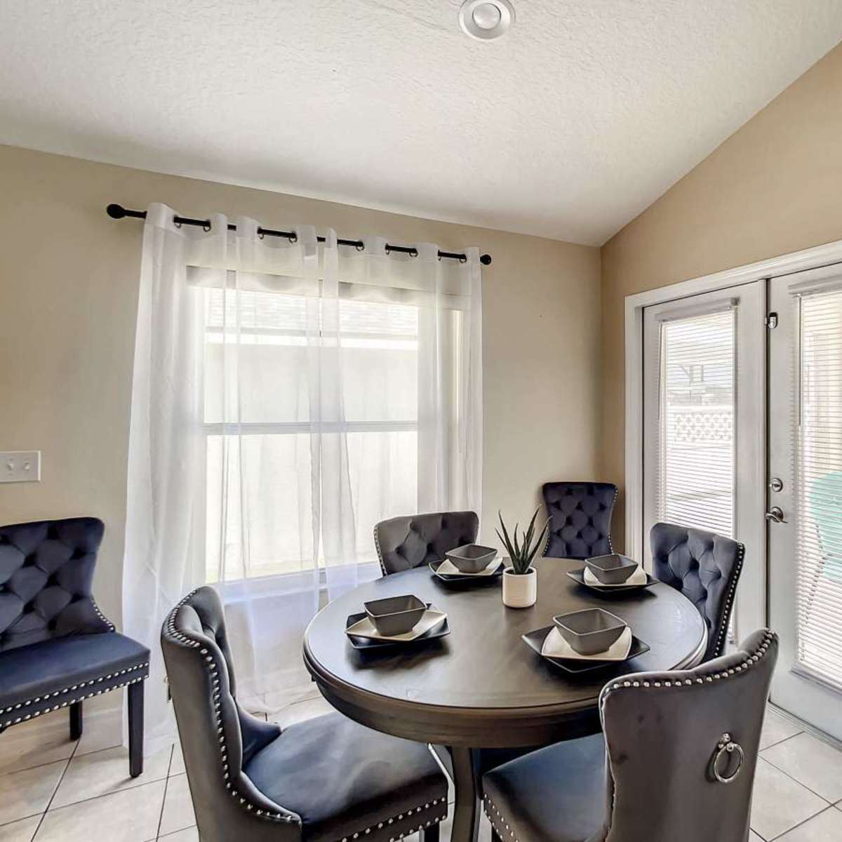 A round dining table is set with four place settings in a small, bright room with a pair of glass doors leading outside.