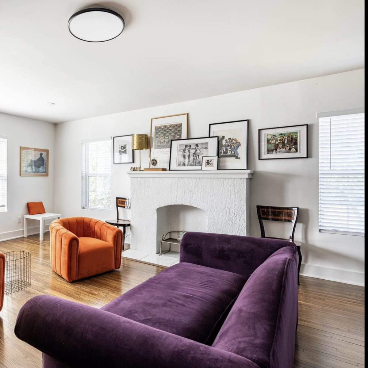 A living room with a purple sofa, two orange armchairs, a wooden dining table, and a white fireplace surrounded by framed pictures.