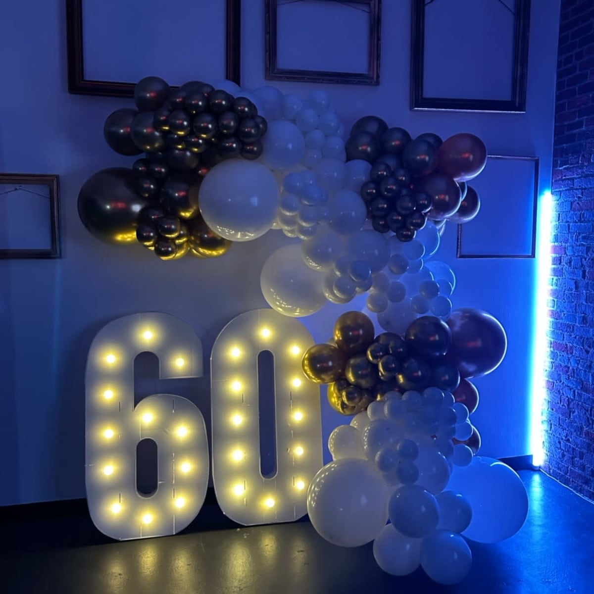 A decorative display of balloons arranged around the illuminated numbers "60" against a wall with empty picture frames.