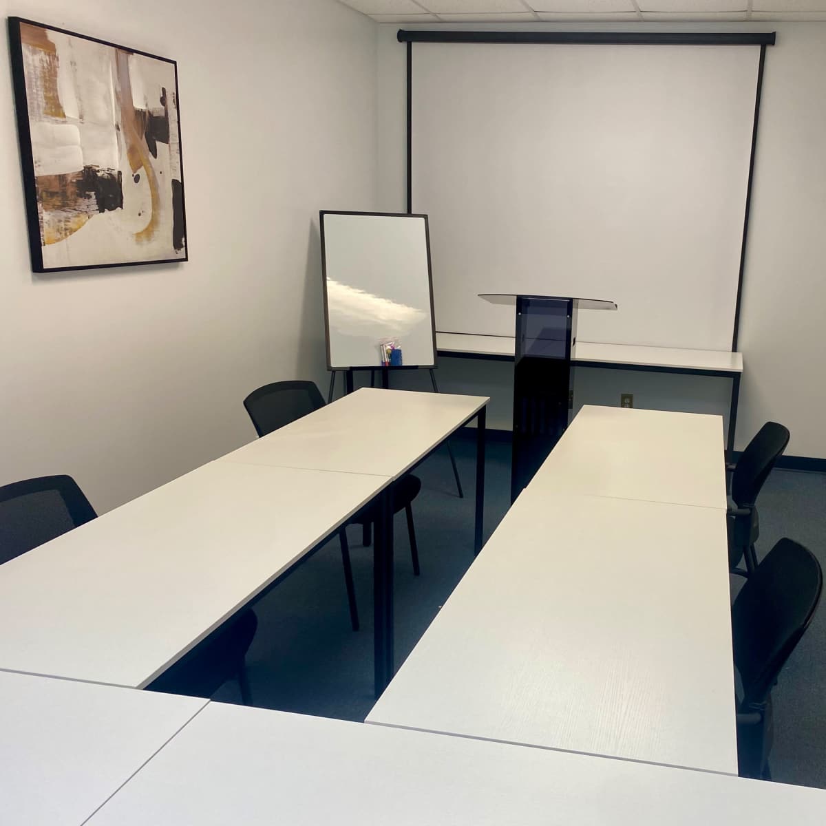 A sparsely furnished meeting room with two long white tables, a podium at the front, a mirror, and a blank projector screen.