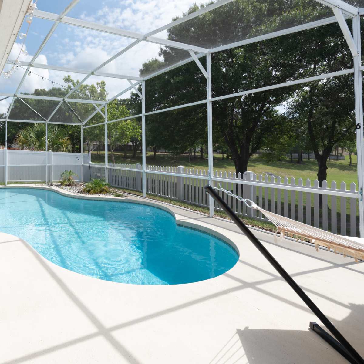 A screened-in patio area with a swimming pool surrounded by greenery and a white fence.