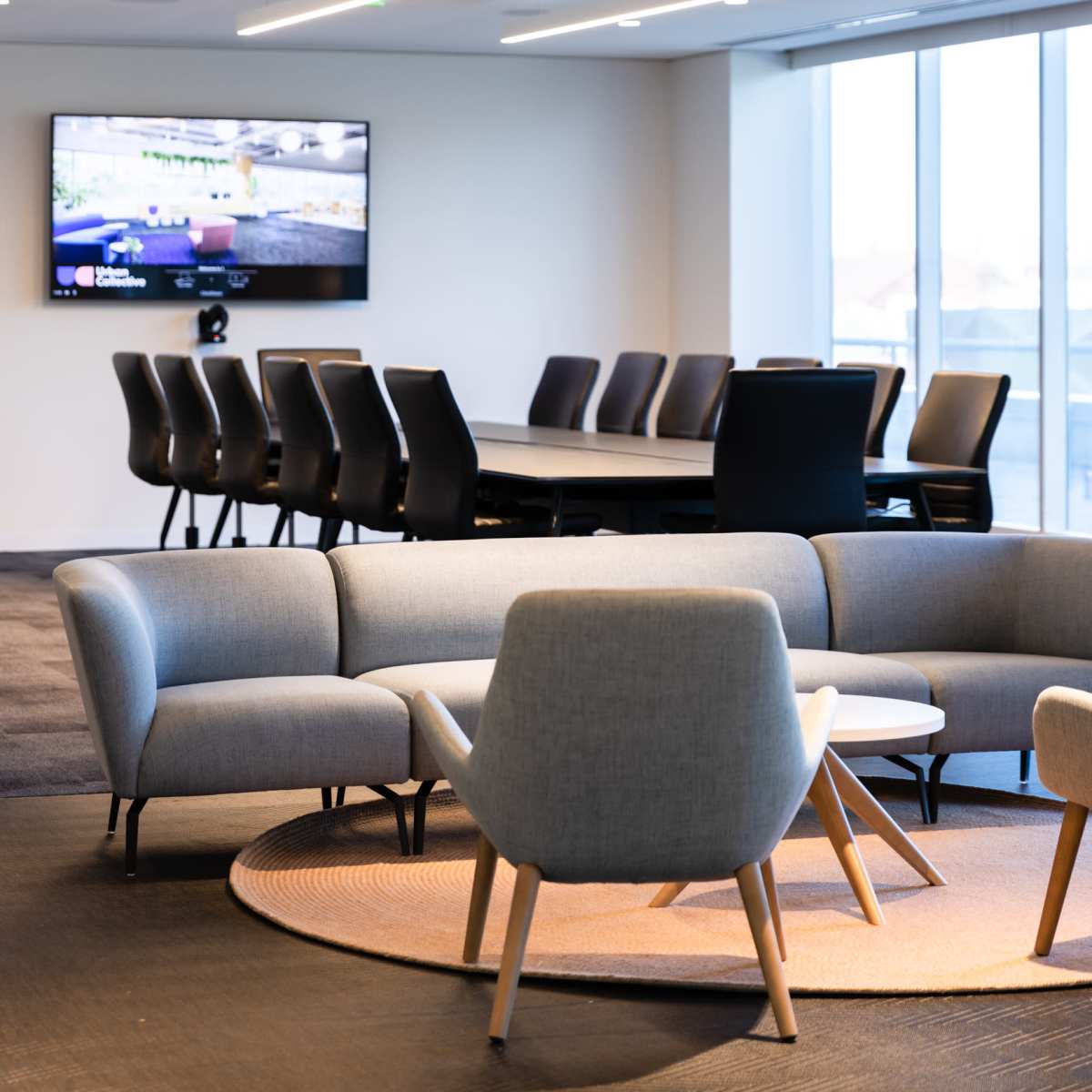 A modern office space featuring a gray sofa set on a circular rug, a large conference table in the background, and round overhead lights illuminating the area.
