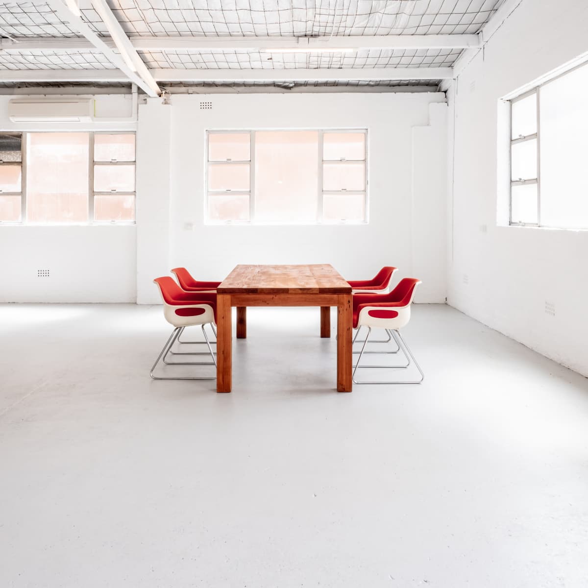 A minimalist meeting room features a wooden table surrounded by four red chairs, with large windows letting in natural light.