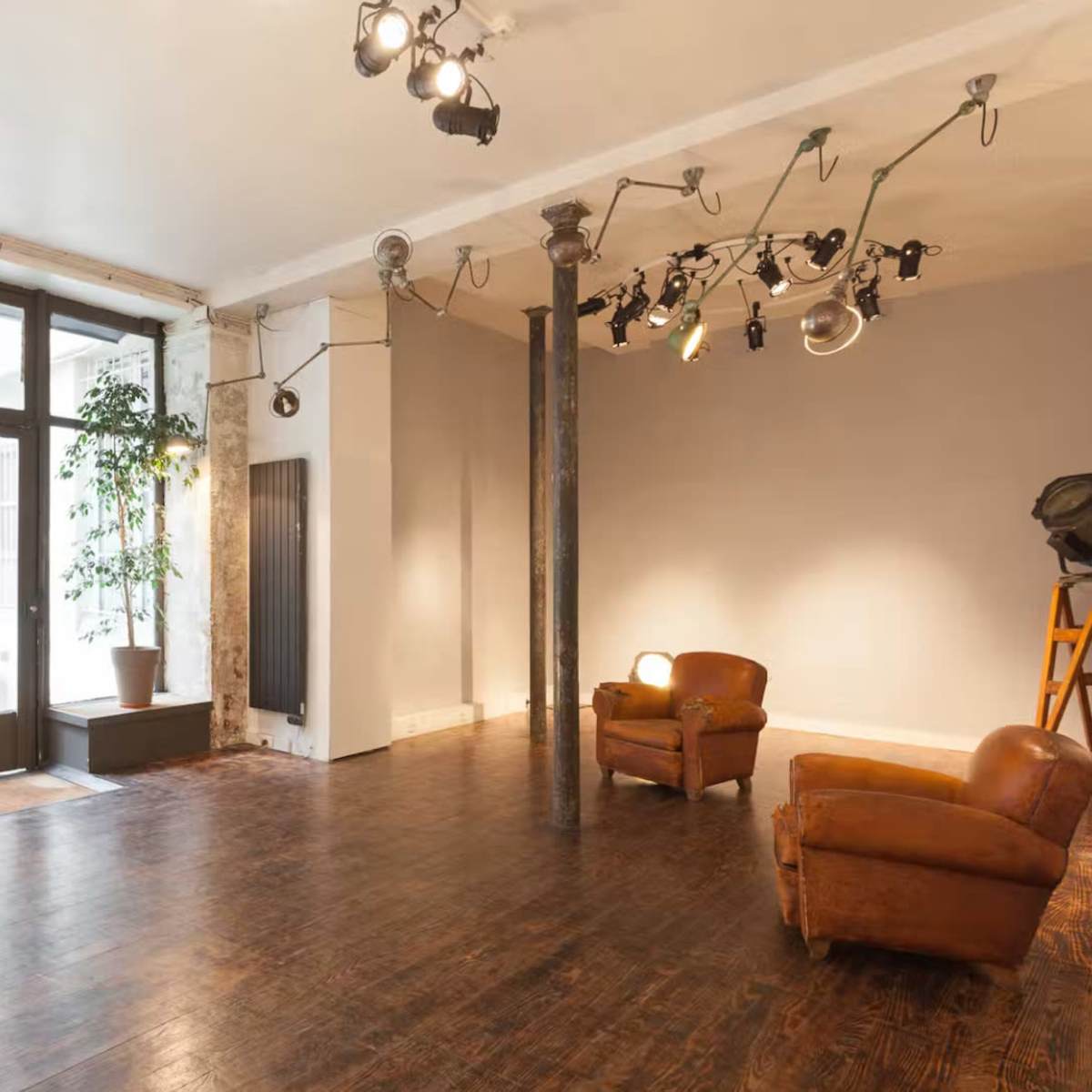 A spacious room with wooden flooring, two brown leather chairs, large windows, and a lighting setup on the ceiling.