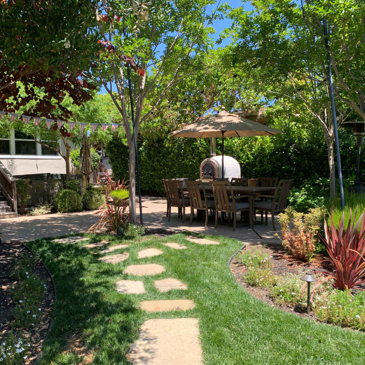 A landscaped backyard features a stone pathway leading to a dining area under a patio umbrella, surrounded by greenery and flowering plants.