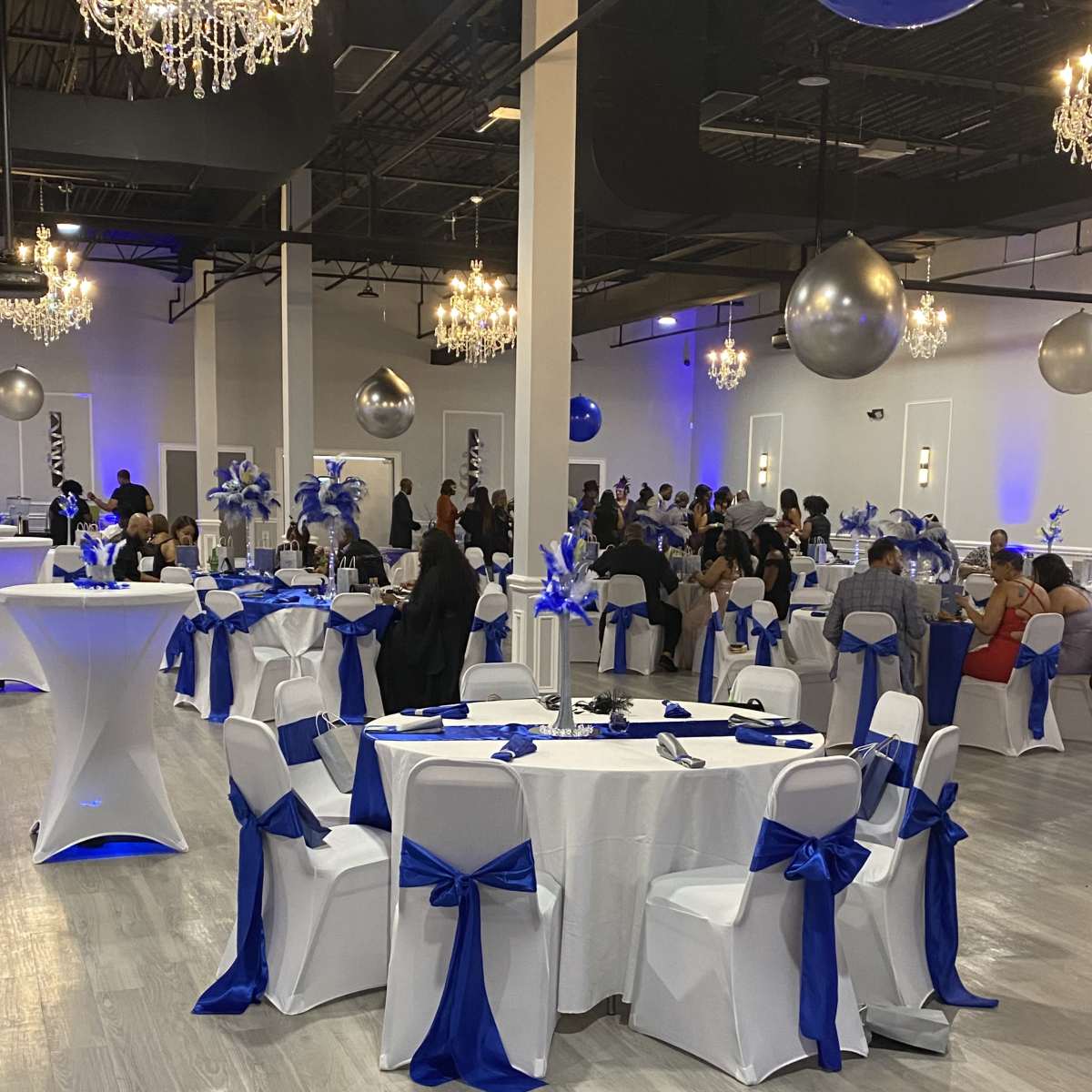 A spacious event venue is set up with white tables adorned with blue satin bows, featuring chandeliers and large silver decor elements hanging from the ceiling, while guests mingle throughout the room.