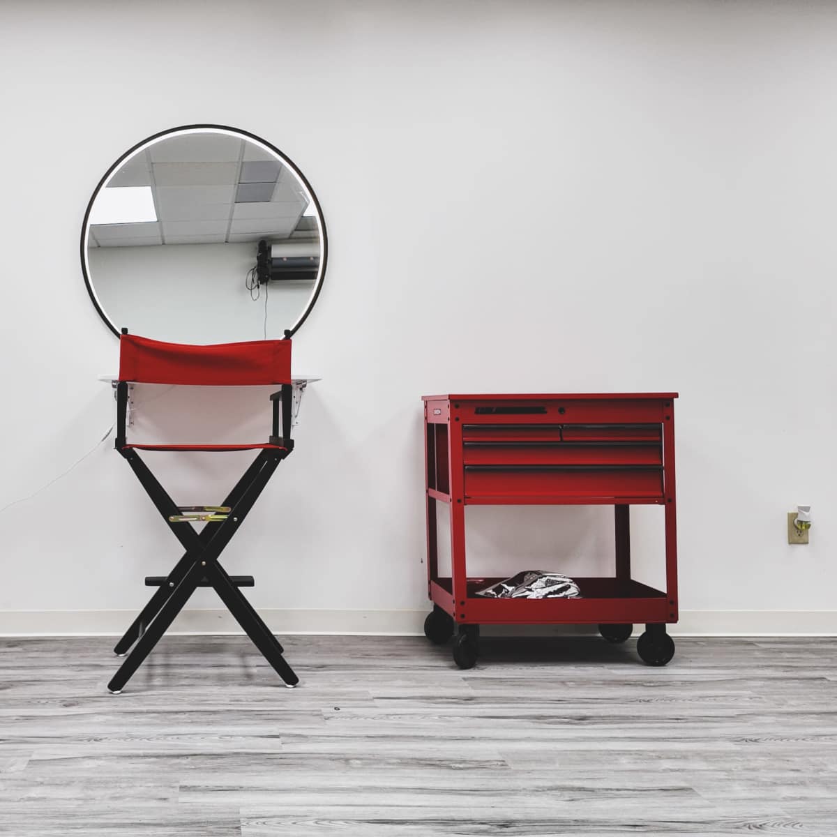 A circular mirror hangs above a foldable chair next to a red rolling cart in a minimalist room with white walls and wooden flooring.