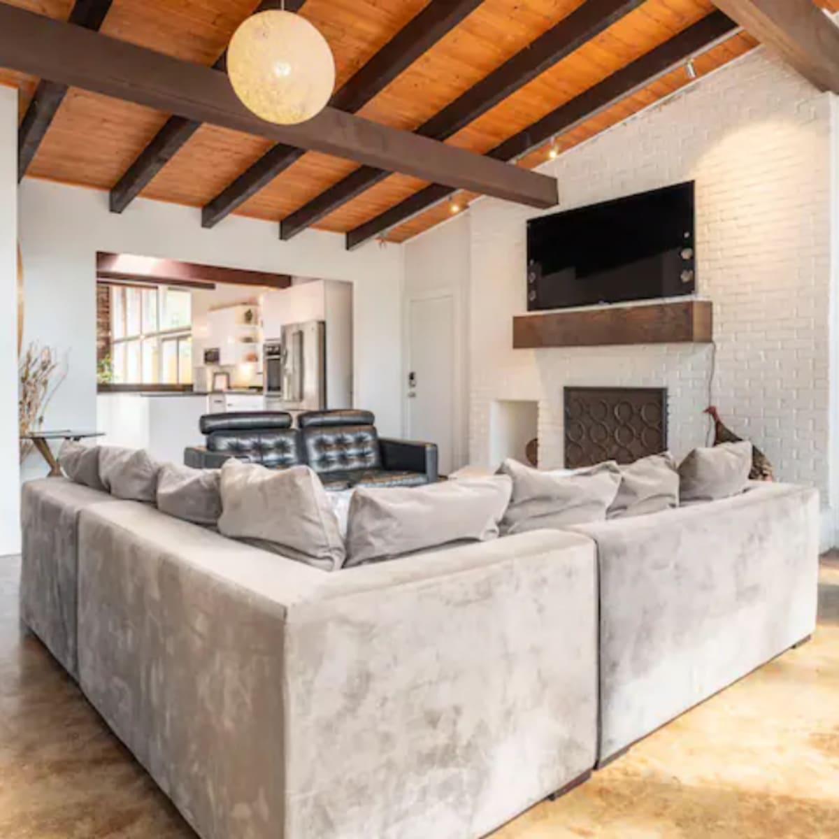 A modern living room featuring a large L-shaped sofa, wooden beams on the ceiling, and a brick fireplace with a mounted television above it.