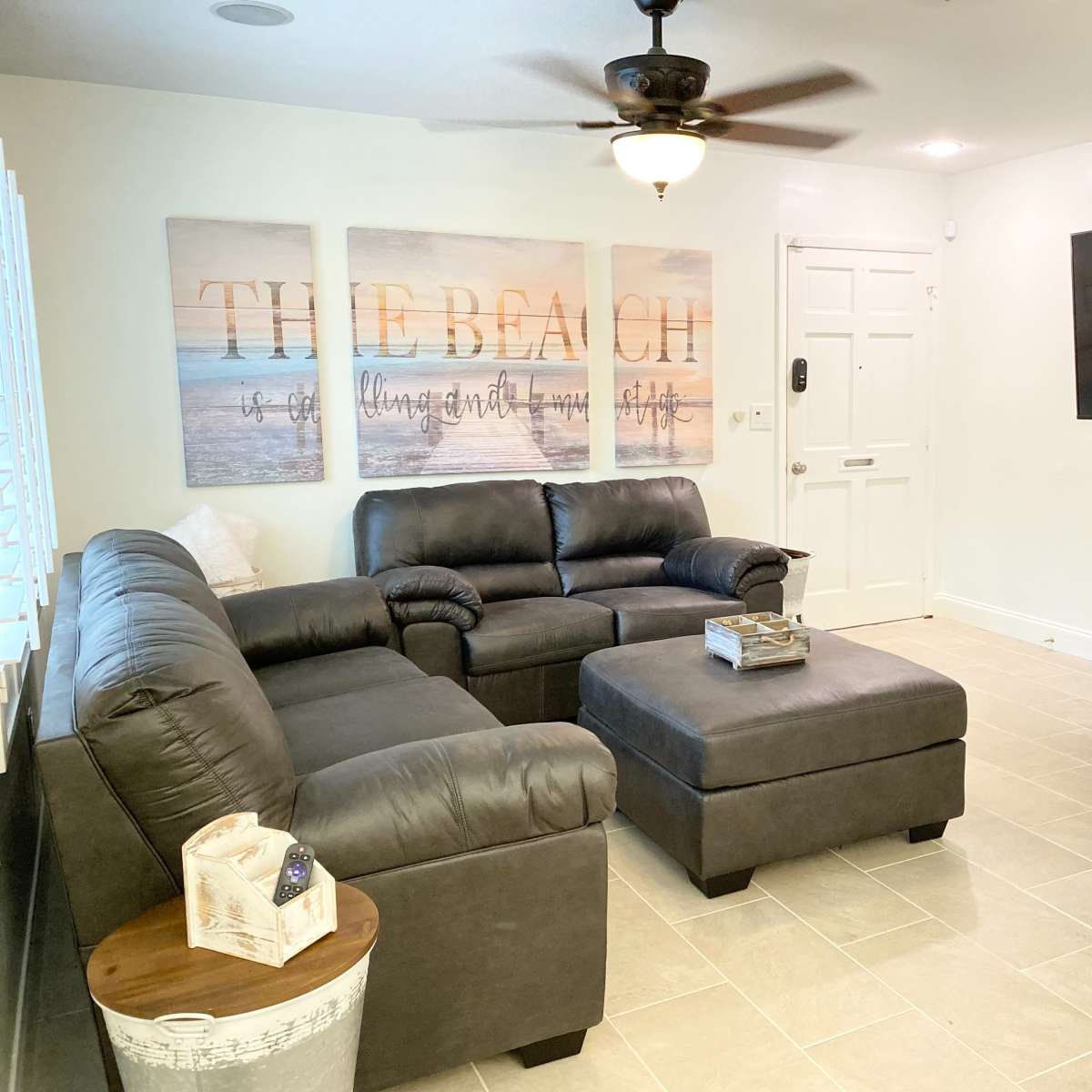 A living room features a black leather sectional sofa, an ottoman, a large television mounted on the wall, and decor that includes canvas prints with beach-themed text.