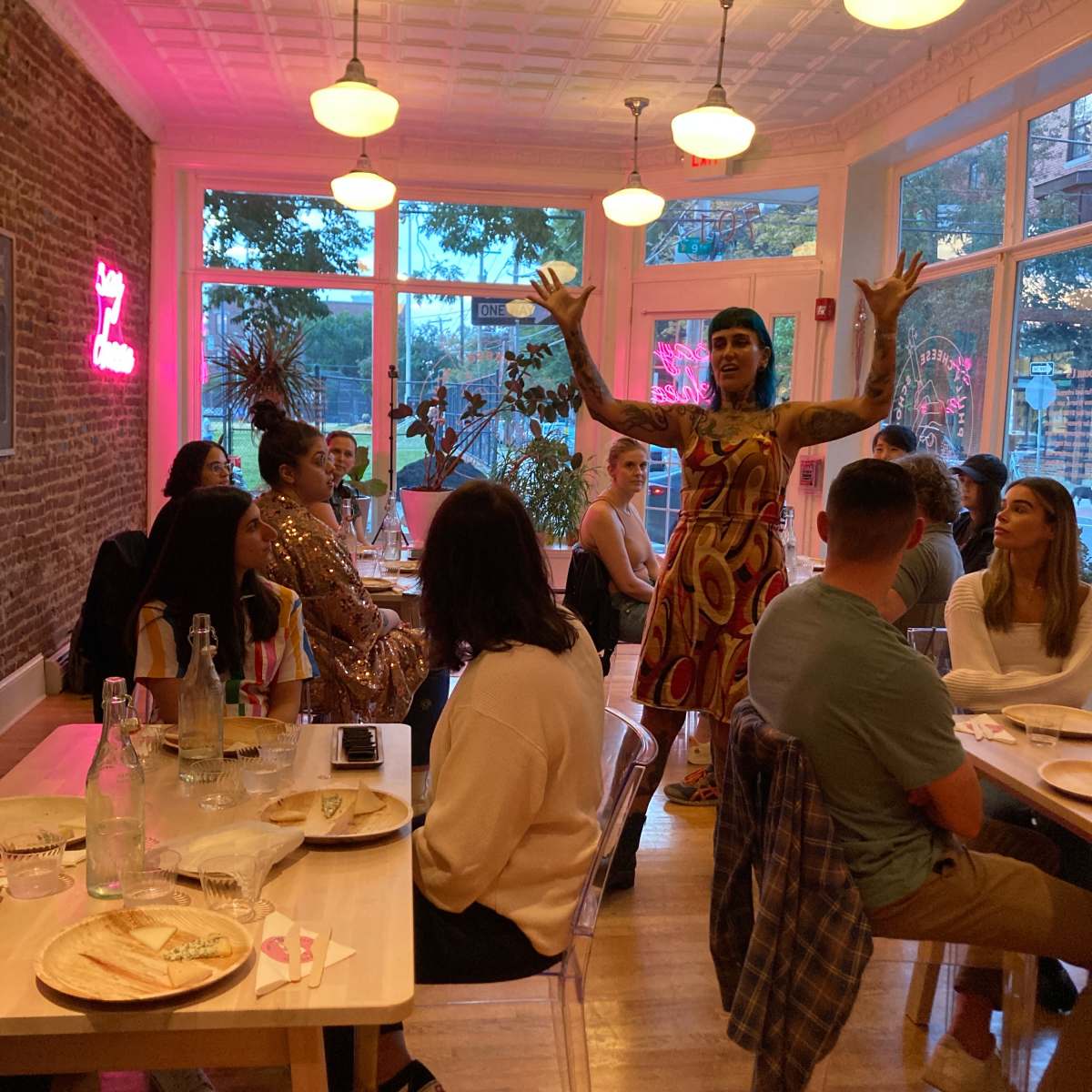 A performer in a colorful dress stands with arms raised in a brightly lit restaurant while guests sit at tables eating.