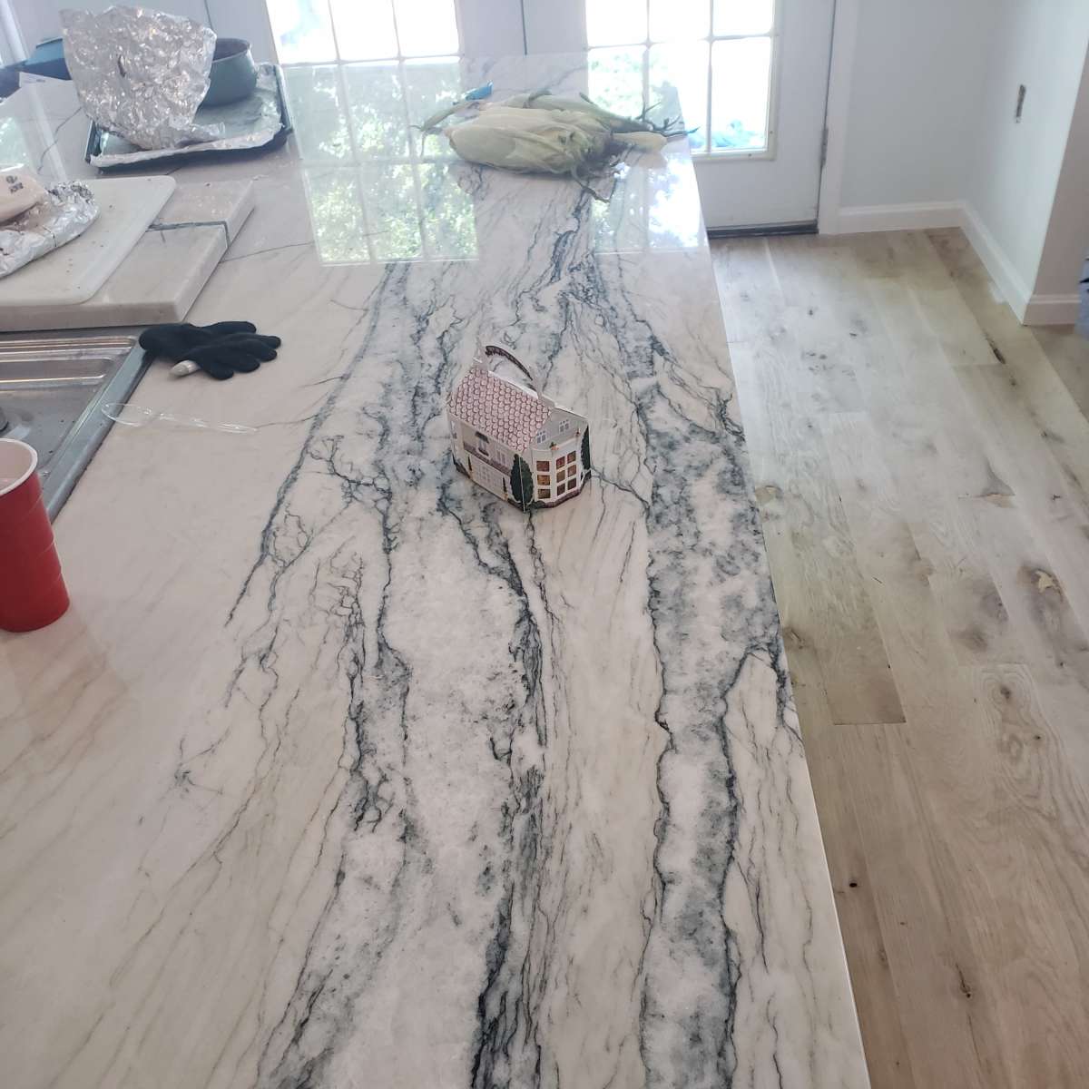 A marble countertop with a swirling black and white pattern displays a small toy house on its surface, alongside a red cup and a piece of green produce.