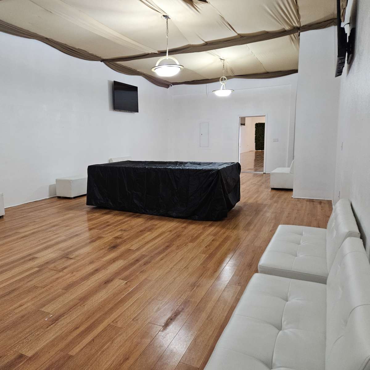 The room features a wooden floor with white walls, two light fixtures, a black-covered table in the center, and several white sofas arranged around the space.