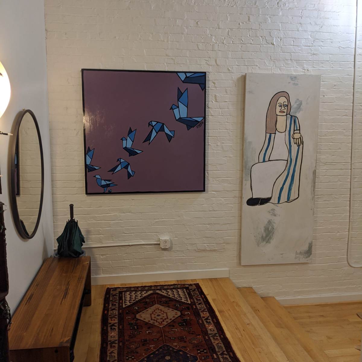 A corner of a room featuring two abstract paintings on the white brick wall, a round mirror, a wooden bench, an umbrella, and a patterned area rug.