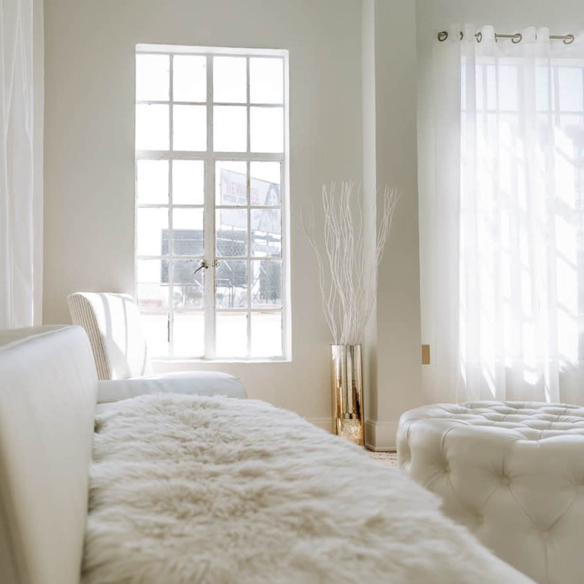 A bright room features a white couch, a tufted ottoman, sheer curtains, and a tall vase with branches near a window.