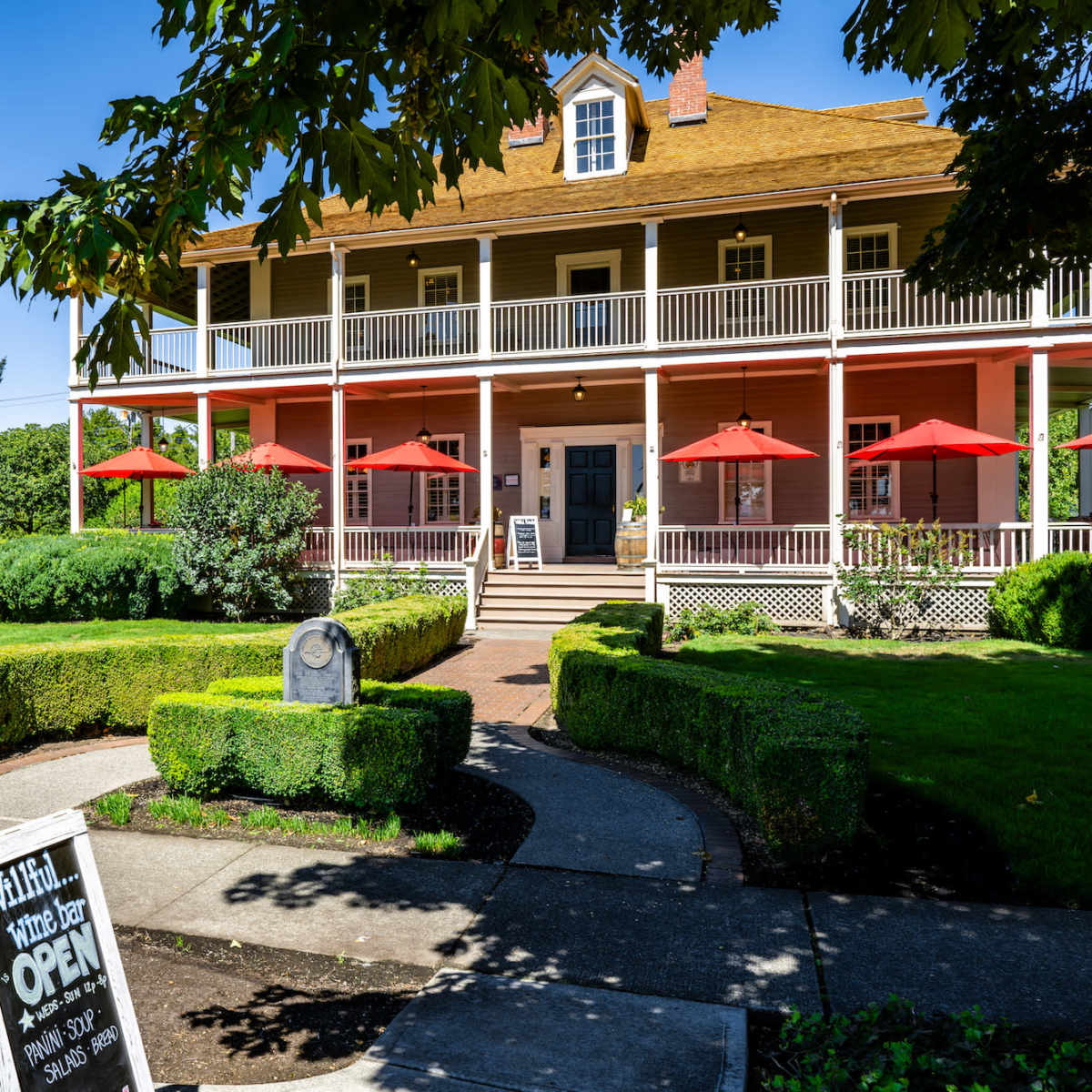 A two-story pink building with a large porch and red umbrellas is surrounded by well-maintained greenery and shrubs.
