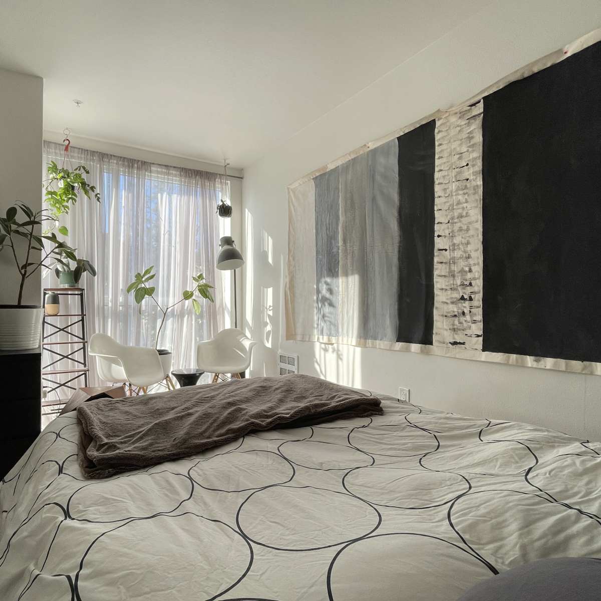 A bedroom features a bed with a patterned duvet, a television stand, and large windows with potted plants, leading to a small seating area with white chairs.