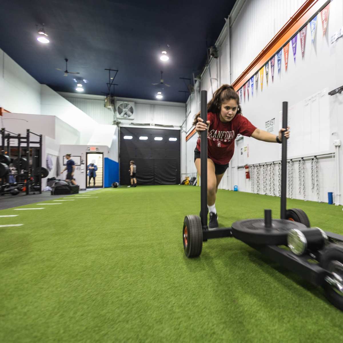 Two athletes are pushing sleds across a turf training area in a gym.