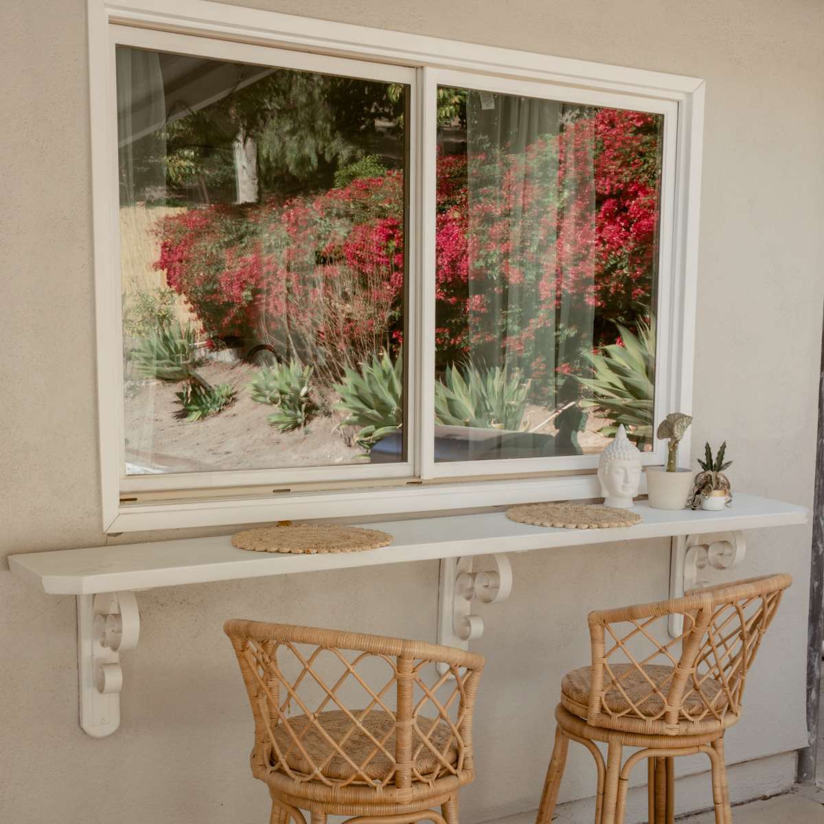 A white countertop with two woven chairs is positioned beneath a large window overlooking a garden with vibrant flowers.