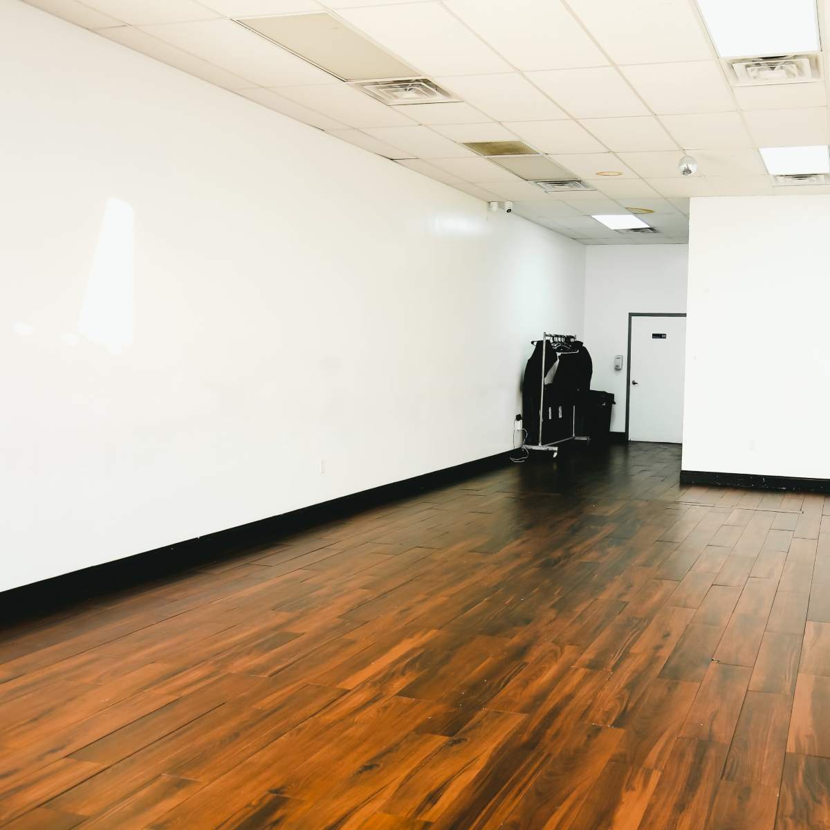 A spacious, empty room with wooden flooring and white walls, featuring a dark rack in the corner.
