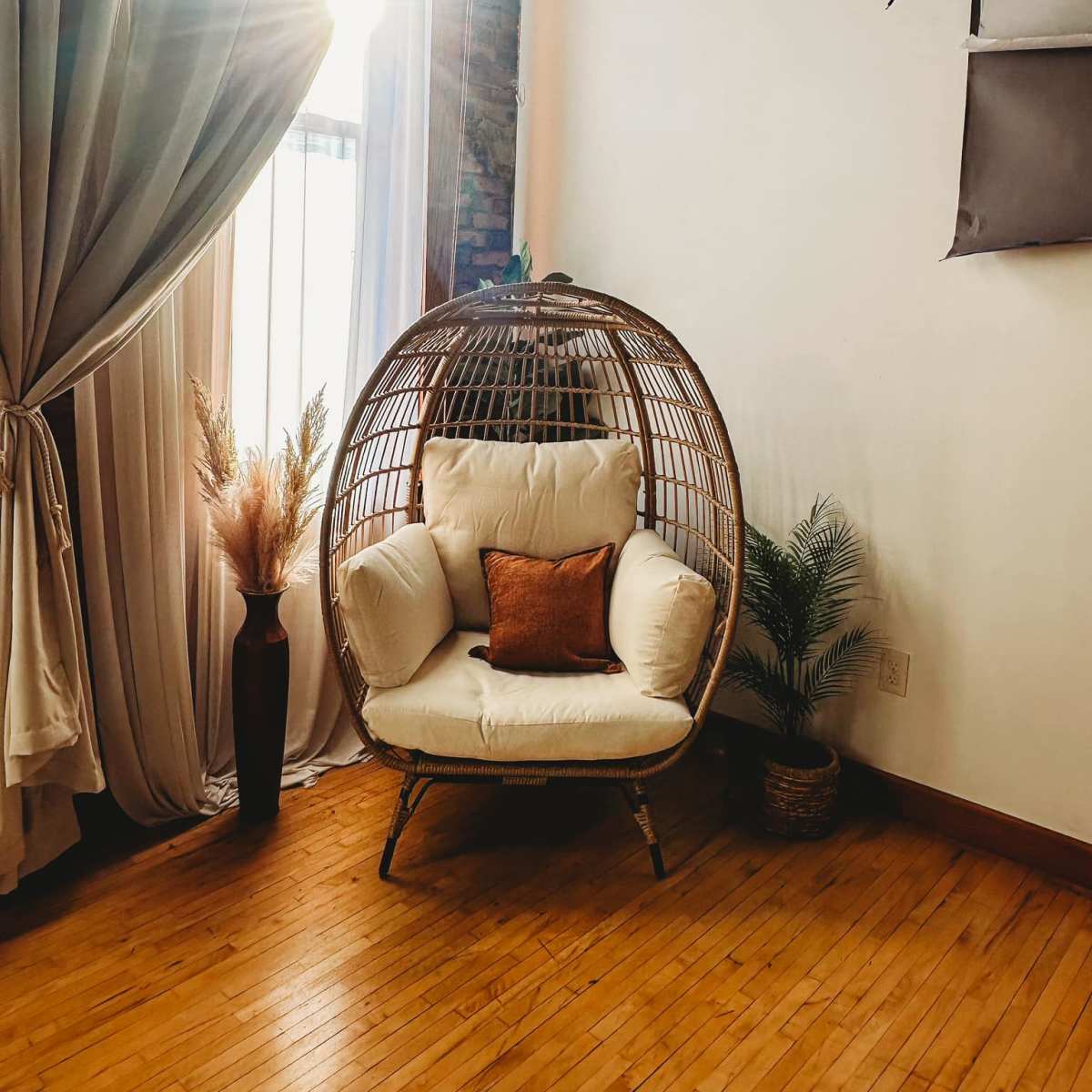 A wicker hanging chair with a cream cushion and a decorative orange pillow is positioned near a large window, surrounded by drapes and plants.