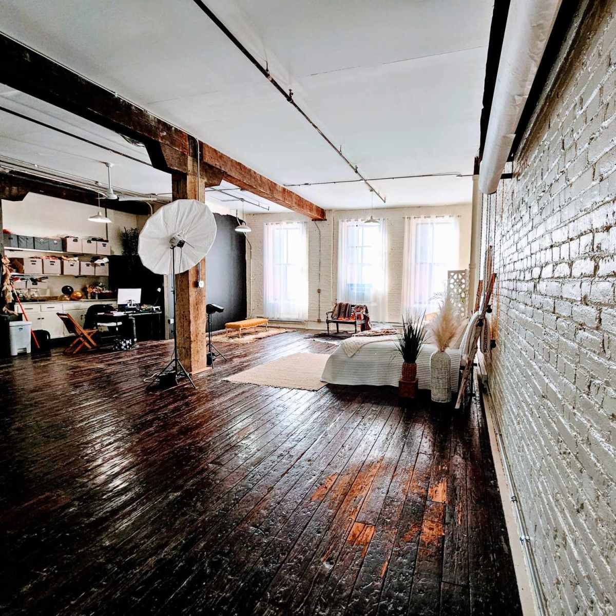 A spacious, well-lit studio with wooden floors, exposed beams, and a sectional area featuring a bed, plants, and a photography umbrella.