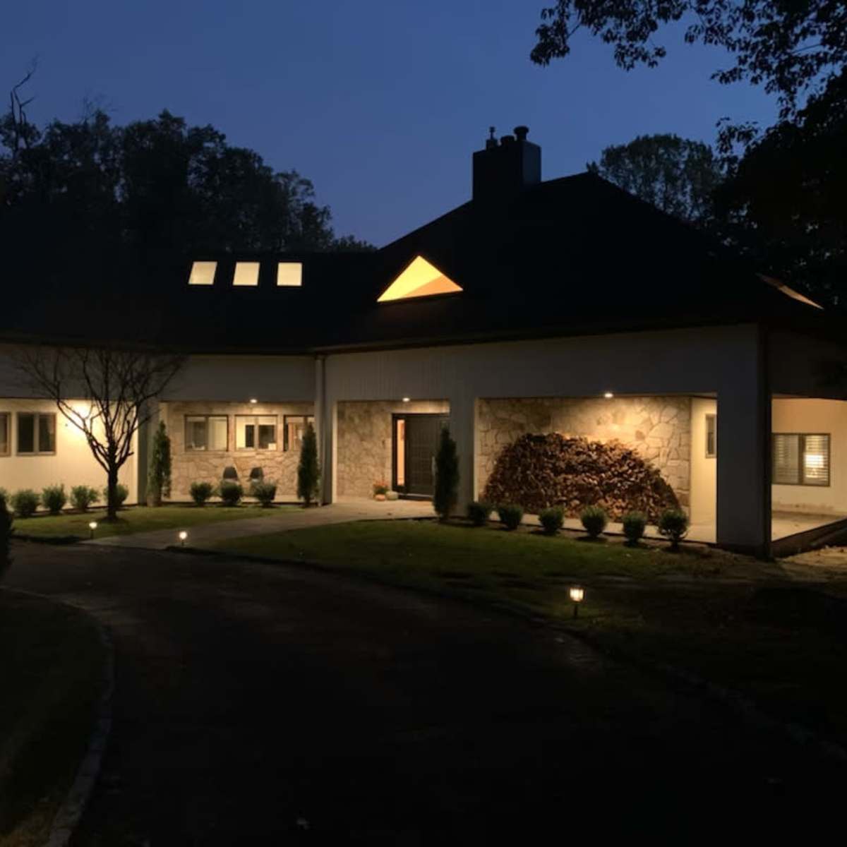 A modern house with stone and white walls is illuminated at night, featuring a landscaped front yard and a curved driveway.