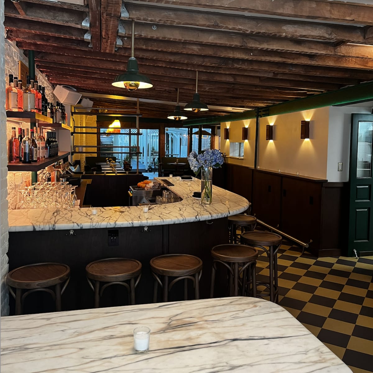 A bar area with a marble countertop, wooden stools, and exposed ceiling beams, featuring shelves of bottles in the background.