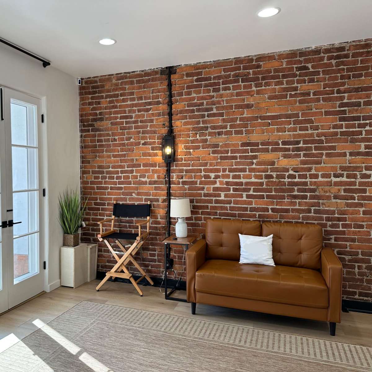 A cozy interior featuring a brown leather sofa, a director's chair, a small plant, and a brick wall with large glass doors.